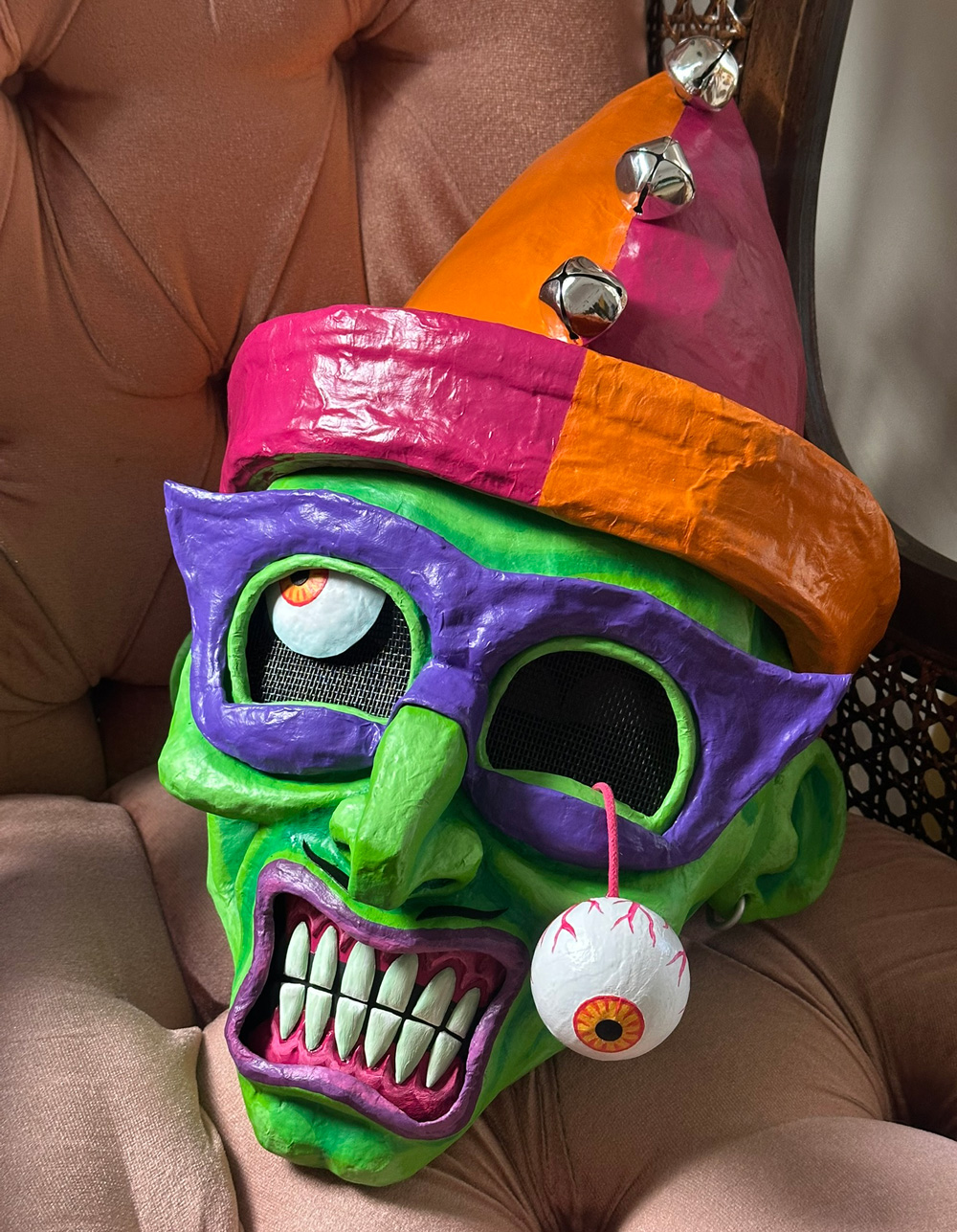 Finished mask; green face, orange and magenta hat, purple eye mask, purple lips, light green teeth, pink gums, and a dangling eyeball with an orange iris.