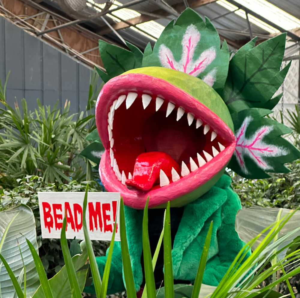 Marjorie wearing her Audrey II mask; a giant venus fly trap head with red lips and sharp teeth; she's standing in a plant store surrounded by plants.