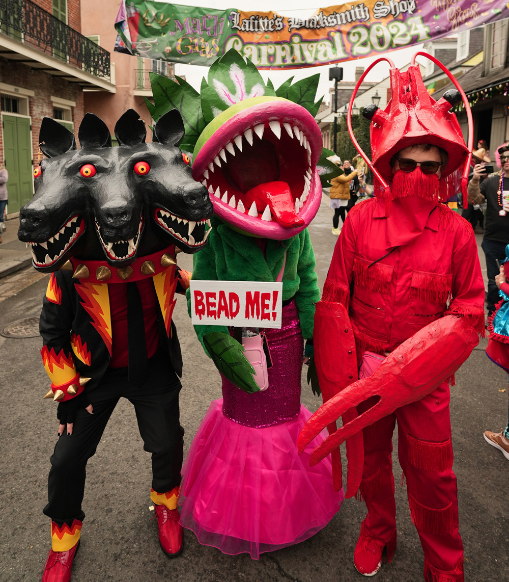 Three costumed people standing in the street: me in a black suit and a three-headed dog mask, Marjorie in her Audrey II mask with a green jacket and pink skirt, and a guy in a bright red crawfish costume.