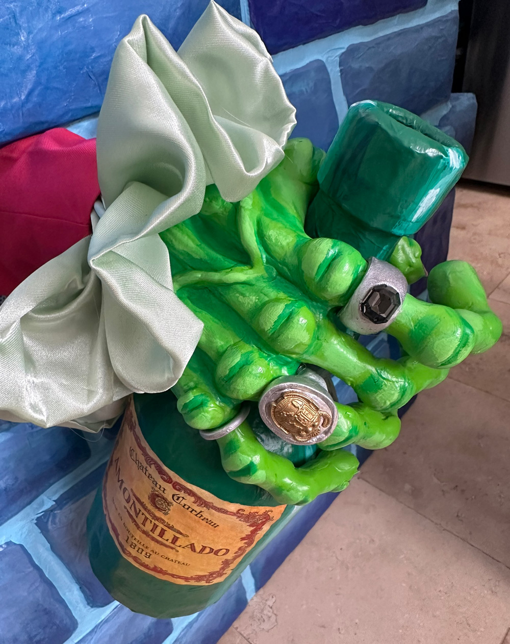 A zombie hand holding a bottle and poking through a hole in a blue brick wall