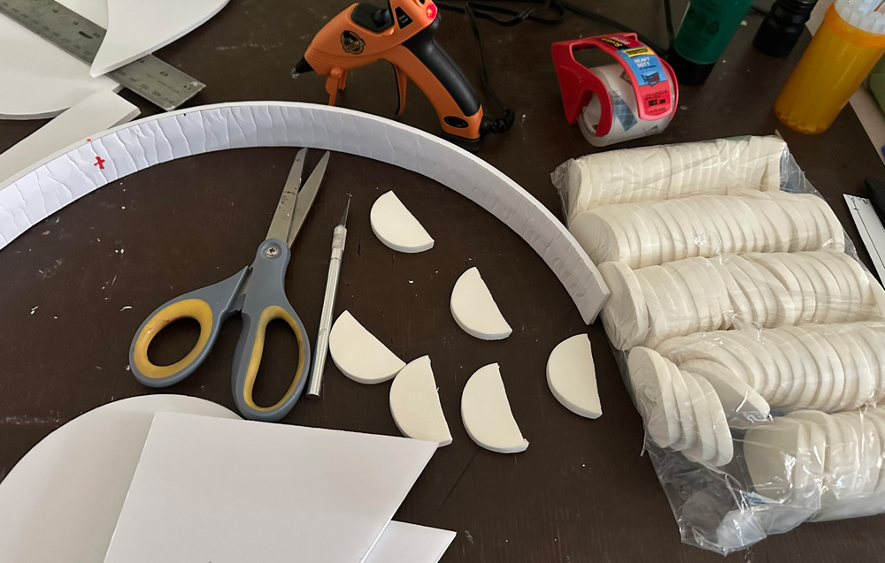 Some items scattered on a table: a thin strip of foam board, a hot glue gun, and some small circular makeup pads that have been cut in half.