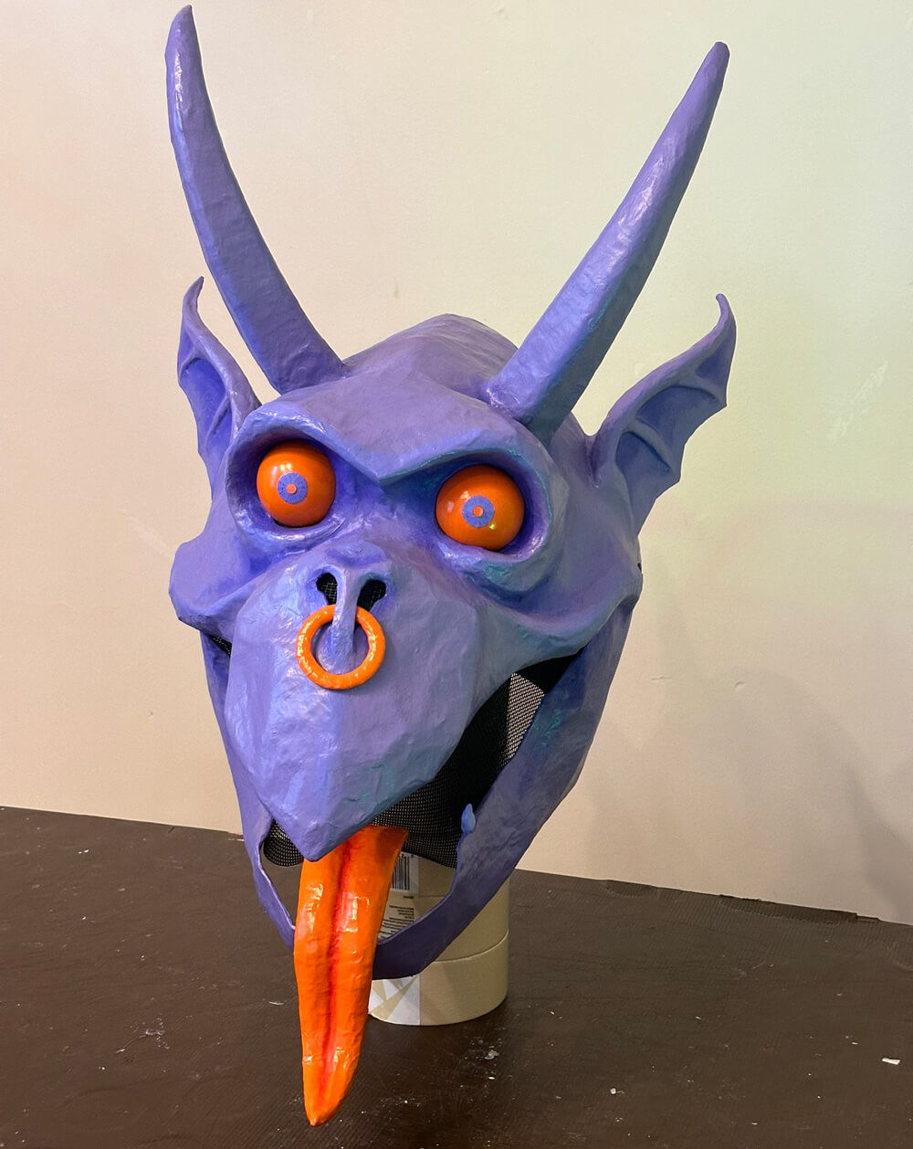 Front view of the finished gargoyle mask, painted purple, with orange eyes, an orange nose ring, and orange tongue.