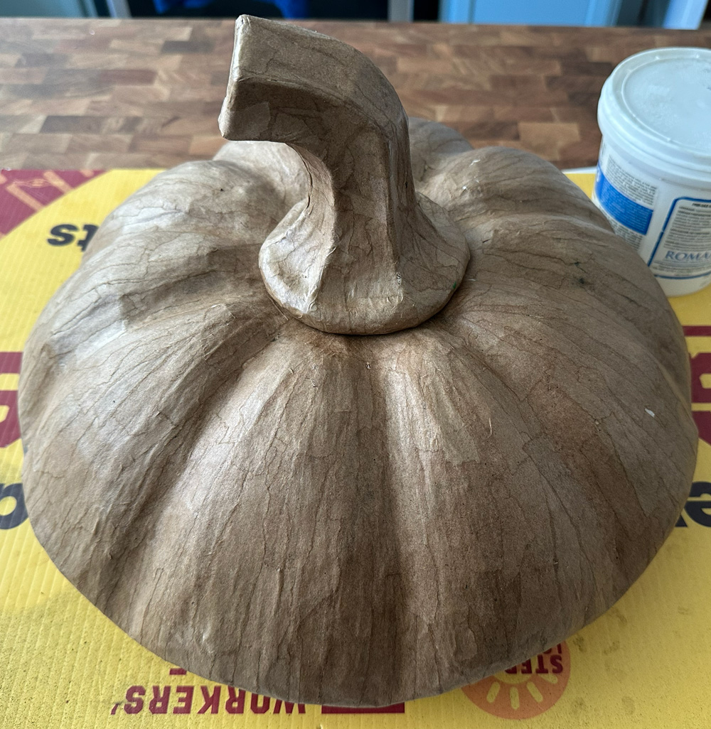 My paper mache pumpkin lid hat sculpture, unpainted, just brown paper. It's a slightly overhead view. The shapes of the small strips of paper mache are visible but they're laying very smooth.