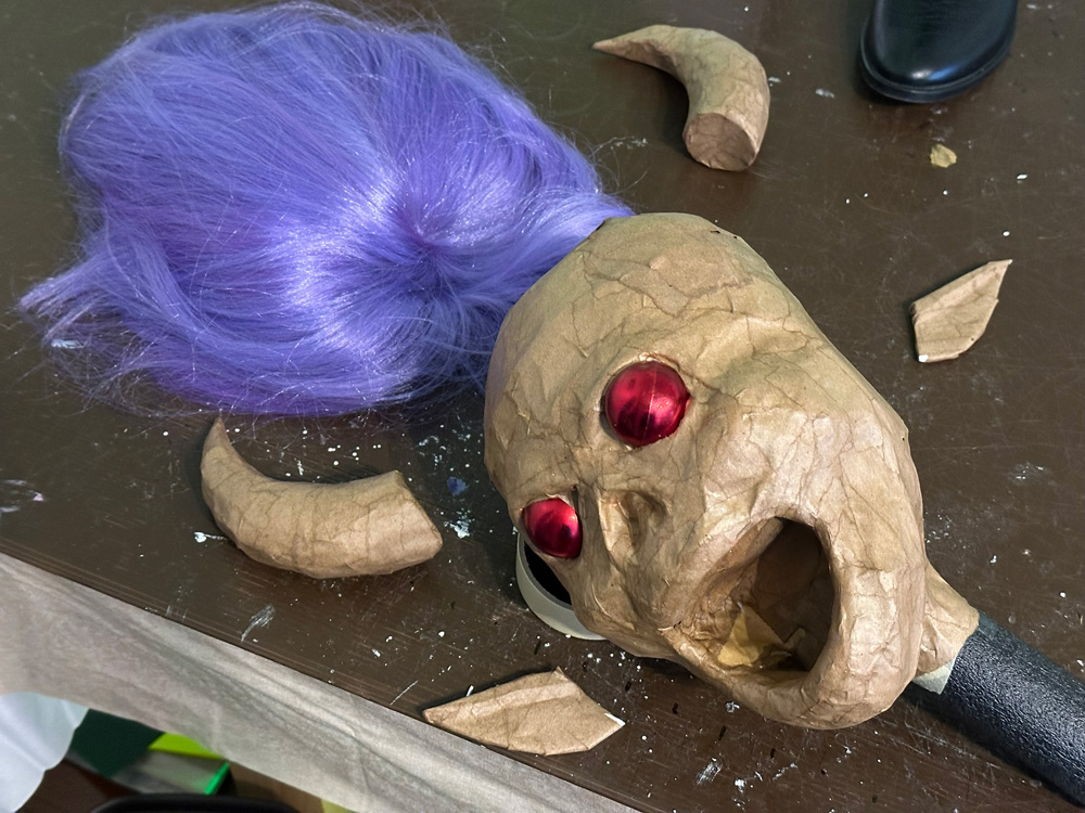 The parts of the demon lady's head all lying on a table; a purple wig, and the rest of the pieces are brown paper mache: two horns, two ears, and a head, with red Christmas ornaments for eyes