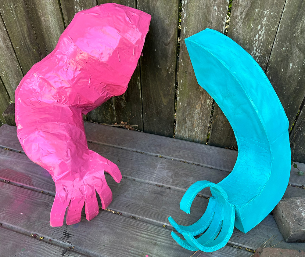 A big pink muscular monster arm, and a blue primitive robot arm, sitting on a table outside