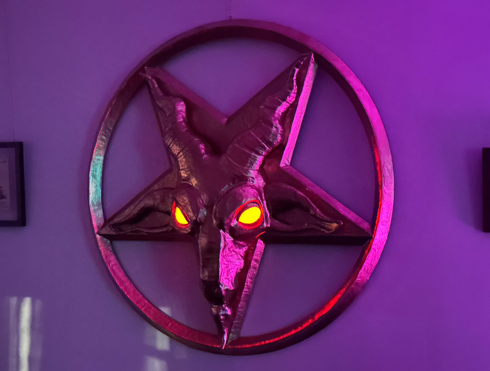 The completed gold pentagram with goat head hanging on my wall at night in dim purple lighting with its eyes lit up looking orange-ish.