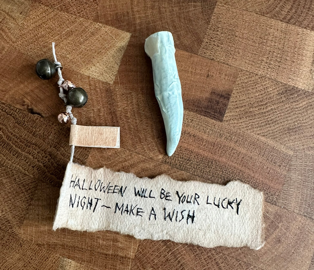 A fake pointy tooth on a table. Next to it is a strip of paper with a fortune written on it; it says, Halloween will be your lucky night; make a wish. The slip of paper is attached to a string, and the string has four tiny jingle bells tied onto it.