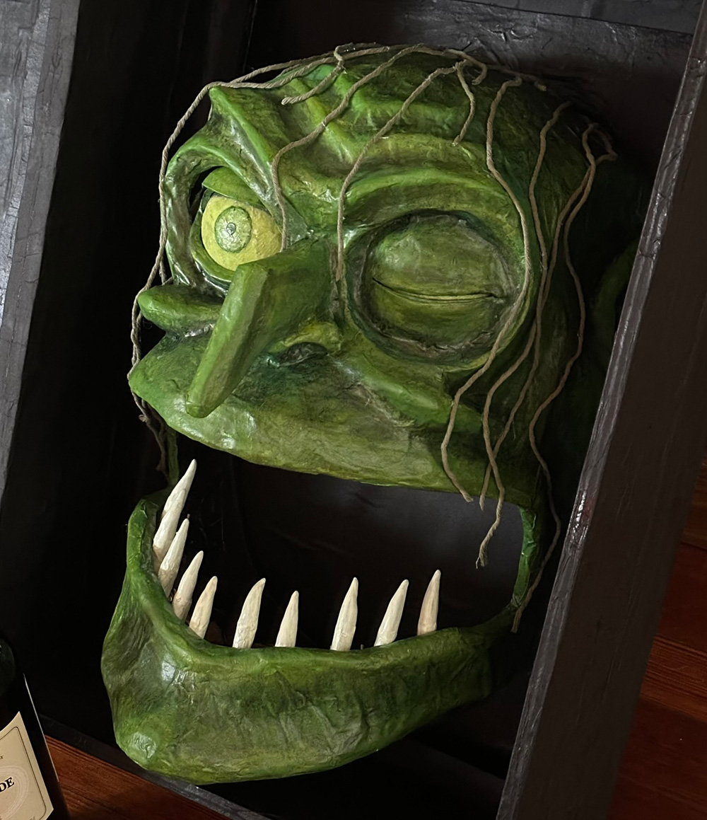The painted green witch head resting in the fake wooden box. She now has one big open eye that's bright yellow-ish green all over.