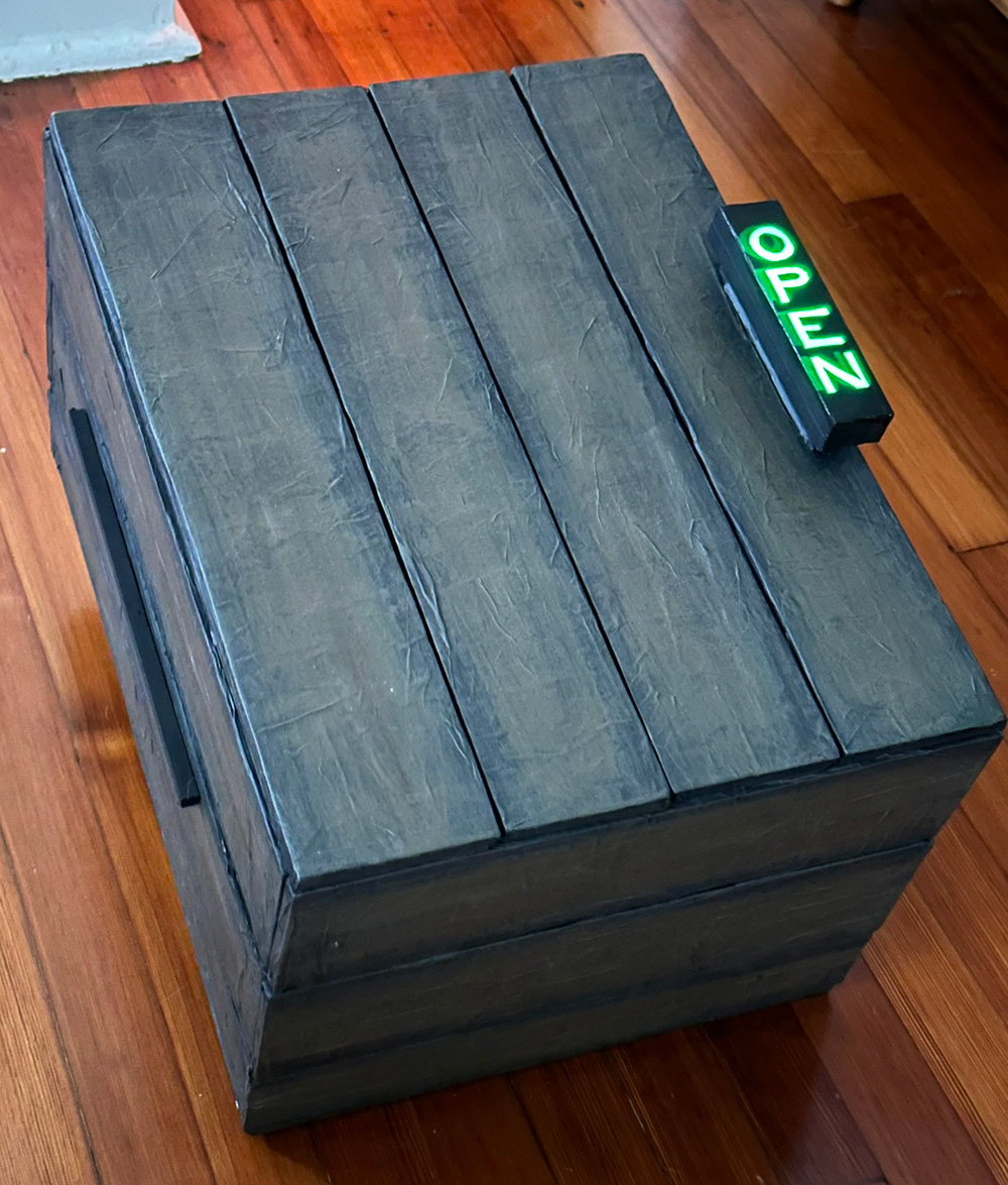 A view of the fake wooden box, closed. It's very dark brown, almost black. The handle has the word open on it, which is glowing green from the lights inside.