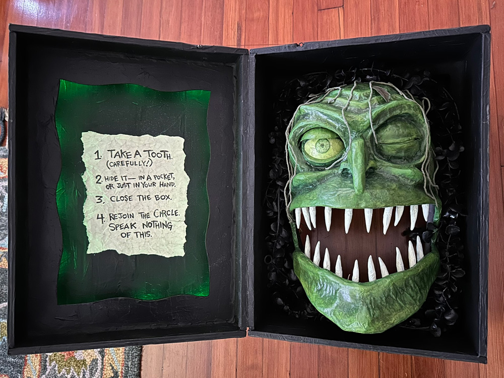 A view of the fake wooden box, open. On the left, inside the lid, there's a piece of old-looking paper with some writing on it. Green recessed light surrounds it. On the right is the witch's face with twenty pointy teeth.