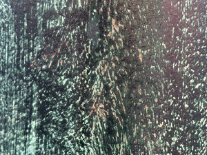 Close up of a scratched metal texture on a light pole which has been painted various colors and all of the paint is very weathered; it's mostly very dark greenish black with some visible spots of lighter green and brown